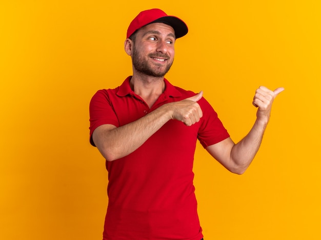 Lächelnder junger kaukasischer Lieferbote in roter Uniform und Mütze, der auf die Seite schaut und Daumen nach oben zeigt, isoliert auf oranger Wand