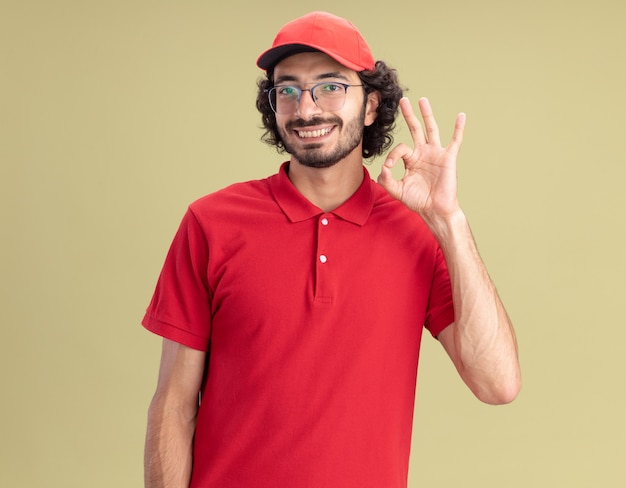 Lächelnder junger kaukasischer Lieferbote in roter Uniform und Mütze mit Brille, der das OK-Zeichen auf olivgrüner Wand isoliert tut