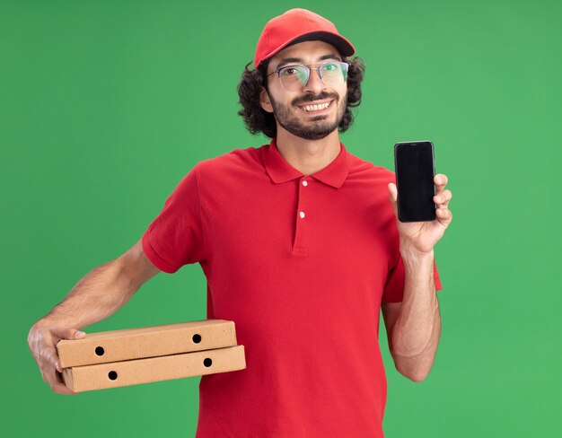 Lächelnder junger kaukasischer Lieferbote in roter Uniform und Mütze mit Brille, die Pizzapakete mit Handy hält