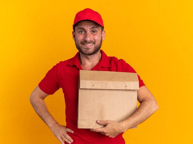 Lächelnder junger kaukasischer Liefermann in roter Uniform und Mütze, der ein Papierpaket hält, das die Hand auf der Taille hält und die Kamera isoliert auf der orangefarbenen Wand betrachtet