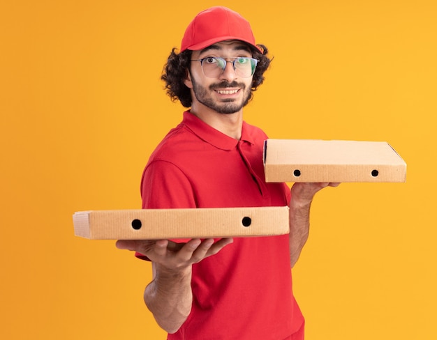 Kostenloses Foto lächelnder junger kaukasischer liefermann in roter uniform und mütze mit brille, der in der profilansicht steht und pizzapakete isoliert auf oranger wand hält und ausstreckt