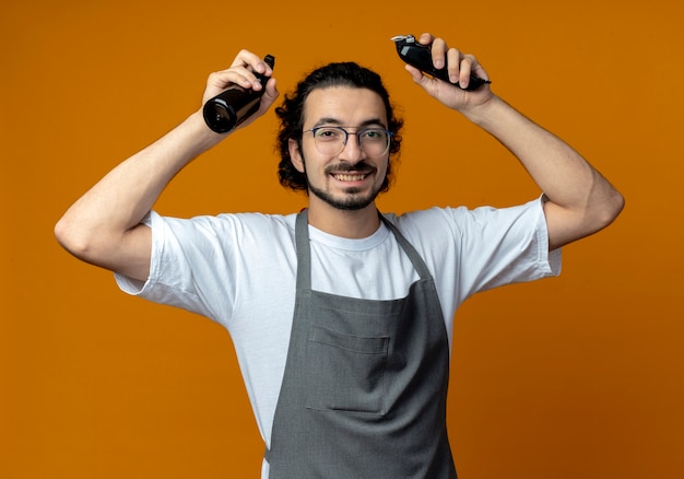 Lächelnder junger kaukasischer männlicher Friseur, der Brille und gewelltes Haarband in der einheitlichen Erhöhung der Sprühflasche und der Haarschneidemaschine lokalisiert auf orangefarbenem Hintergrund trägt