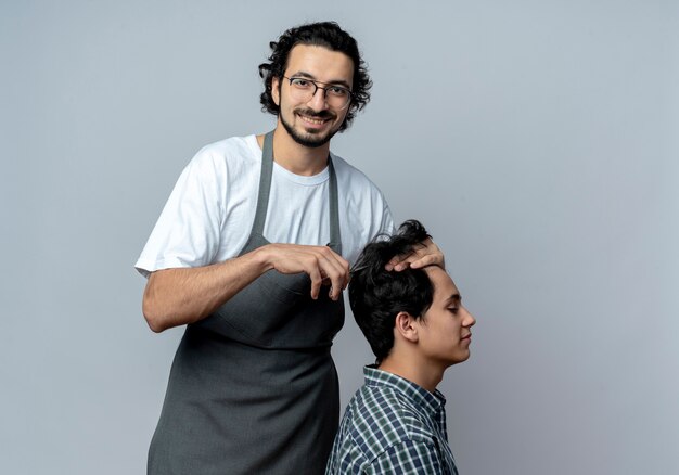 Lächelnder junger kaukasischer männlicher Friseur, der Brille und gewelltes Haarband in Uniform trägt, die Haarschnitt für seinen jungen Klienten lokalisiert auf weißem Hintergrund mit Kopienraum tun