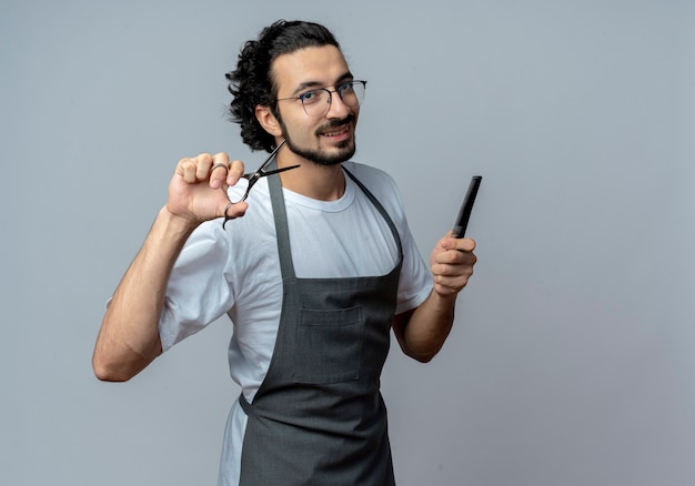 Lächelnder junger kaukasischer männlicher Friseur, der Brille und welliges Haarband in der Uniform hält, die Schere und Kamm hält, lokalisiert auf weißem Hintergrund mit Kopienraum