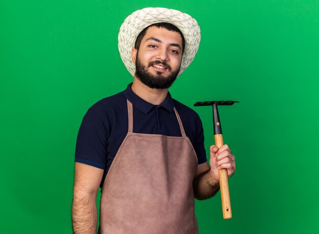 lächelnder junger kaukasischer männlicher Gärtner, der Gartenhut hält, der Rechen lokalisiert auf grüner Wand mit Kopienraum hält