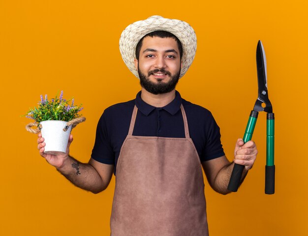 lächelnder junger kaukasischer männlicher Gärtner mit Gartenhut mit Blumentopf und Gartenschere isoliert auf oranger Wand mit Kopierraum with