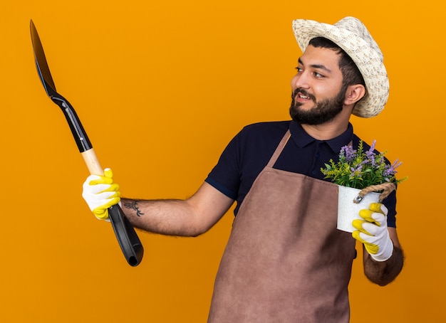 lächelnder junger kaukasischer männlicher Gärtner mit Gartenhut und Handschuhen, der Blumentopf hält und Spaten einzeln auf oranger Wand mit Kopierraum betrachtet