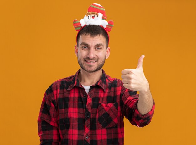 Lächelnder junger kaukasischer Mann, der das Weihnachtsmann-Stirnband trägt, das Kamera betrachtet Daumen oben auf orange Hintergrund lokalisiert