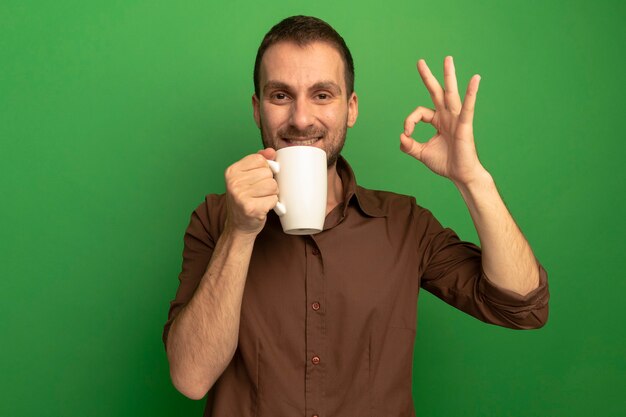 Lächelnder junger kaukasischer Mann, der Tasse Tee hält und Kamera betrachtet, die ok Zeichen lokalisiert auf grünem Hintergrund mit Kopienraum tut