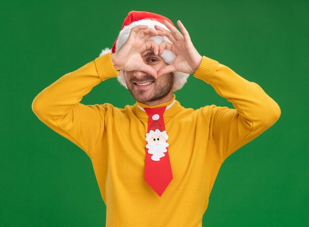 Lächelnder junger kaukasischer Mann, der Weihnachtshut und Krawatte trägt und Kamera betrachtet, die Herzzeichen vor Auge lokalisiert auf grünem Hintergrund tut
