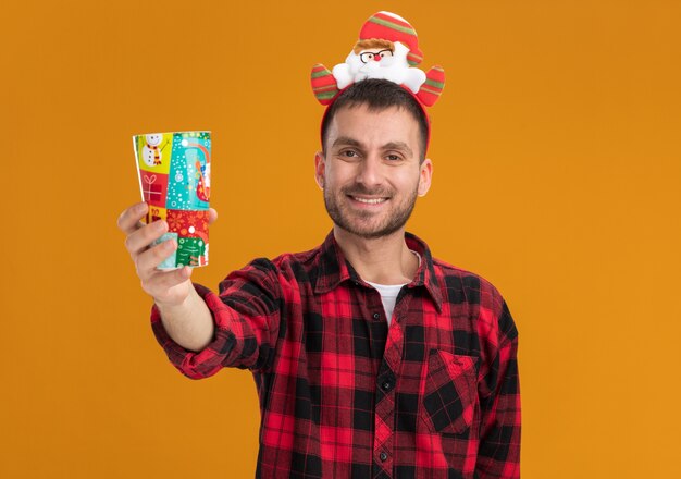 Lächelnder junger kaukasischer Mann, der Weihnachtsmannclaus-Stirnband trägt, das Plastikweihnachtsbecher in Richtung Kamera ausdehnt, Kamera betrachtend Kamera lokalisiert auf orange Hintergrund mit Kopienraum