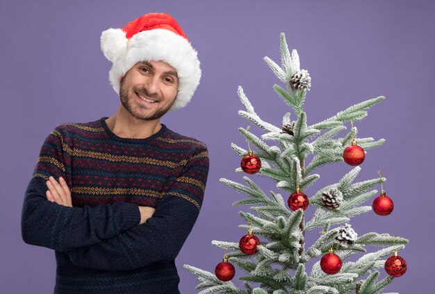 lächelnder junger kaukasischer Mann mit Weihnachtsmütze, der mit geschlossener Haltung in der Nähe des geschmückten Weihnachtsbaums steht und isoliert auf lila Wand aussieht