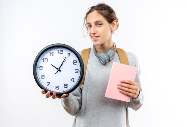 Lächelnder junger Kerl Student mit Rucksack mit Kopfhörern am Hals hält Wanduhr mit Notebook isoliert auf weißer Wand