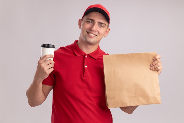 Lächelnder junger lieferbote, der uniform mit kappe hält papiernahrungsmittelpaket mit tasse kaffee lokalisiert auf weißer wand trägt