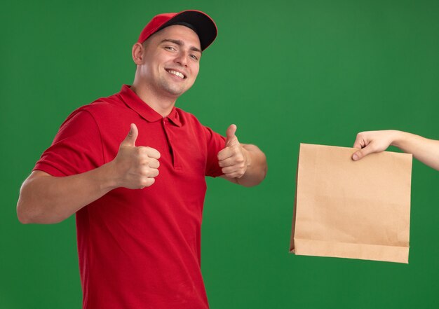 Lächelnder junger Lieferbote, der Uniform und Kappe trägt, die dem Kunden Papiernahrungsmittelpaket geben, das Daumen oben isoliert auf grüner Wand zeigt