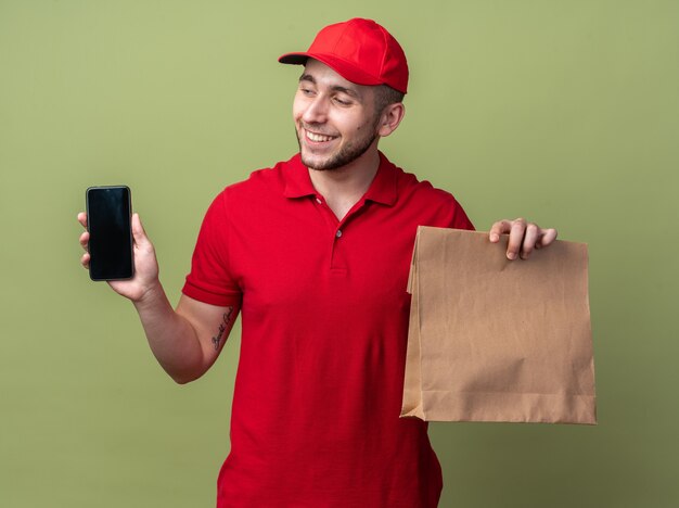 Lächelnder junger Lieferbote in Uniform mit Mütze, der eine Papiertüte mit Lebensmittel hält und in der Hand auf das Telefon schaut