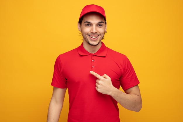 lächelnder junger lieferbote in uniform und kappe, der auf die kamera zeigt, die auf die seite zeigt, die auf gelbem hintergrund isoliert ist