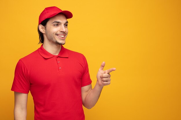 lächelnder junger lieferbote in uniform und kappe, der auf die seite zeigt, die auf gelbem hintergrund mit kopienraum isoliert ist