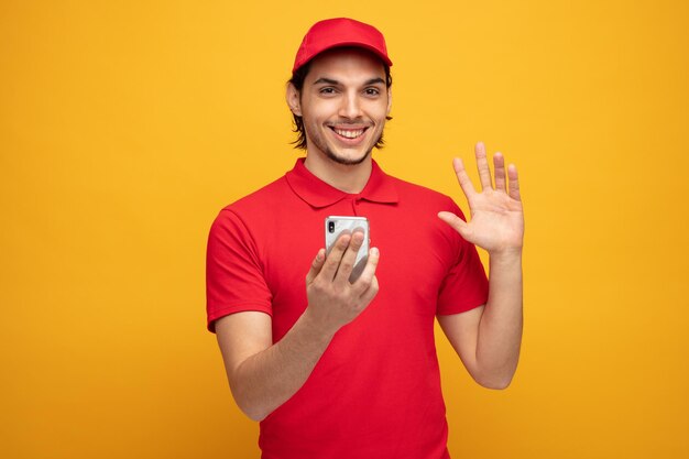 lächelnder junger lieferbote in uniform und kappe, der ein handy hält, das in die kamera schaut, die isoliert auf gelbem hintergrund winkt