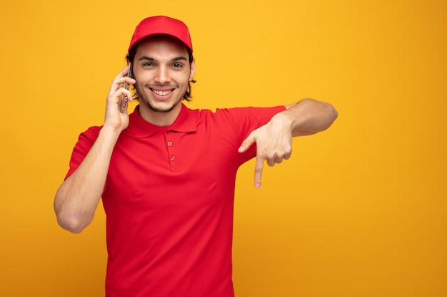 Lächelnder junger Lieferbote in Uniform und Mütze, der in die Kamera blickt, die nach unten zeigt, während er am Telefon spricht, isoliert auf gelbem Hintergrund