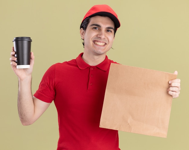 Lächelnder junger Liefermann in roter Uniform und Mütze mit Papierpaket und Plastikkaffeetasse mit Blick auf die Vorderseite isoliert auf olivgrüner Wand