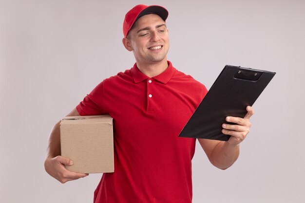Lächelnder junger Liefermann in Uniform mit Mütze, der eine Schachtel hält und die Zwischenablage in seiner Hand isoliert auf weißer Wand betrachtet