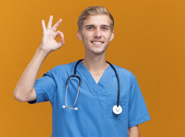 Lächelnder junger männlicher Arzt, der Arztuniform mit Stethoskop trägt, das auf orange Wand lokalisierte Geste zeigt