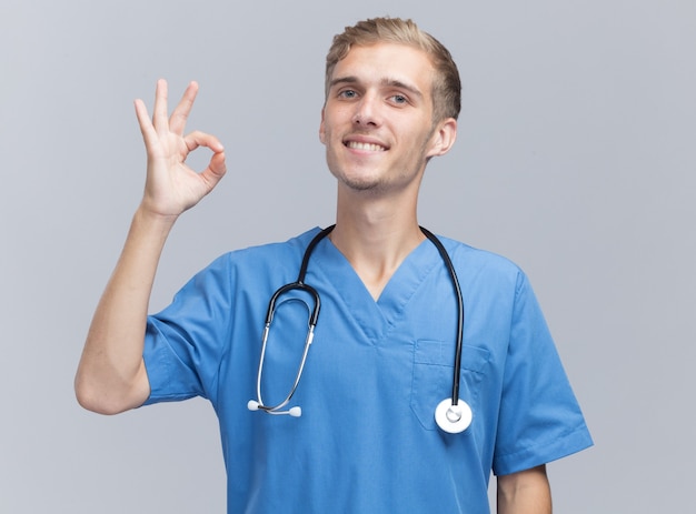 Lächelnder junger männlicher Arzt, der Arztuniform mit Stethoskop trägt, das okay Geste zeigt, die auf weißer Wand lokalisiert wird