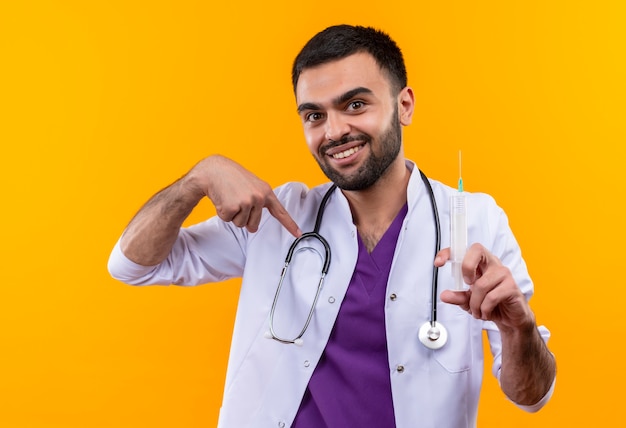 Lächelnder junger männlicher Arzt, der das medizinische Kleid des Stethoskops hält, das Spritze hält, zeigt auf lokalisiertem gelbem Hintergrund
