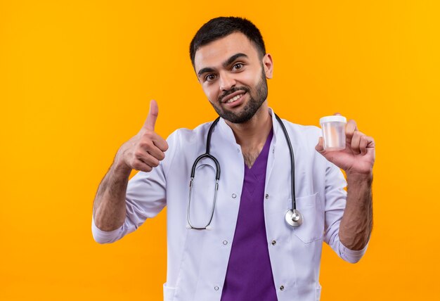 Lächelnder junger männlicher Arzt, der das medizinische Kleid des Stethoskops trägt, das leer hält, kann seinen Daumen oben auf lokalisiertem gelbem Hintergrund
