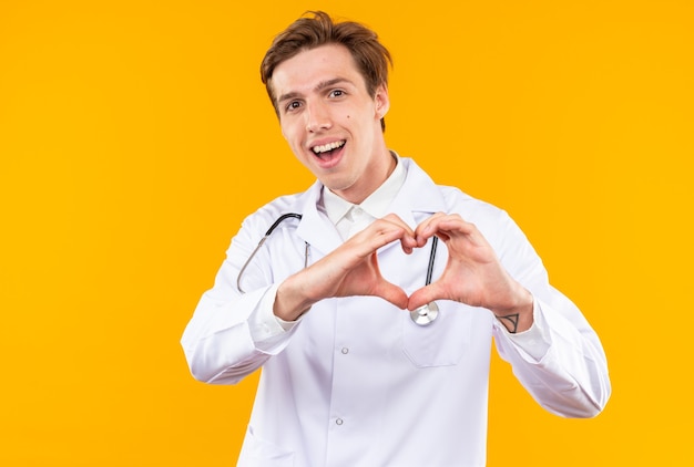 Lächelnder junger männlicher Arzt, der ein medizinisches Gewand mit Stethoskop trägt und eine Herzgeste zeigt, die auf oranger Wand isoliert ist?