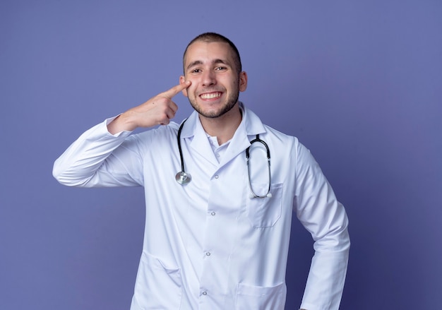 Lächelnder junger männlicher Arzt, der medizinische Robe und Stethoskop trägt, die Finger auf Wange lokalisiert auf lila Wand setzen