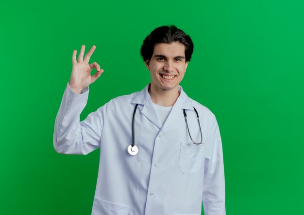 Lächelnder junger männlicher Arzt, der medizinische Robe und Stethoskop trägt, die ok Zeichen lokalisiert auf grüner Wand mit Kopienraum tun
