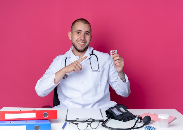 Lächelnder junger männlicher Arzt, der medizinische Robe und Stethoskop trägt, sitzt am Schreibtisch mit Arbeitswerkzeugen, die auf Packung der Kapseln lokalisiert auf rosa Wand halten und zeigen