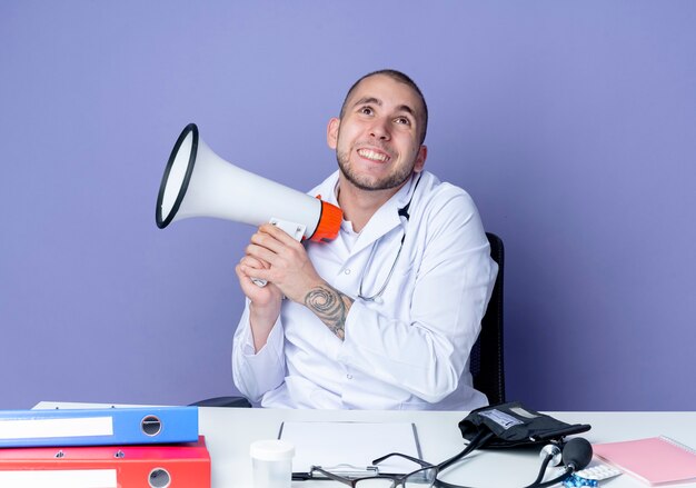 Lächelnder junger männlicher Arzt, der medizinische Robe und Stethoskop trägt, sitzt am Schreibtisch mit Arbeitswerkzeugen, die Lautsprecher halten und lokalisiert auf lila Wand suchen