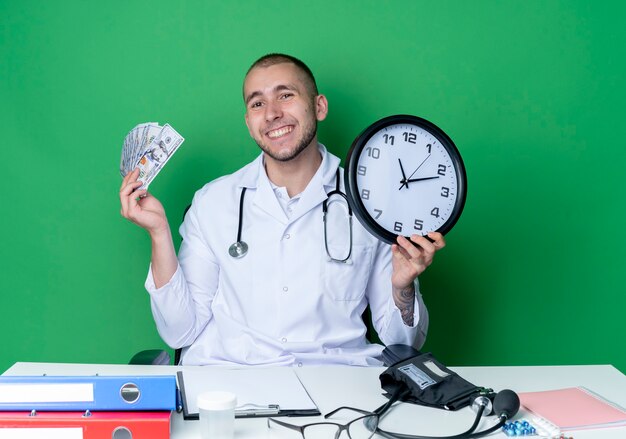 Lächelnder junger männlicher Arzt, der medizinische Robe und Stethoskop trägt, sitzt am Schreibtisch mit Arbeitswerkzeugen, die Uhr und Geld lokalisiert auf grüner Wand halten
