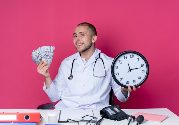 Lächelnder junger männlicher Arzt, der medizinische Robe und Stethoskop trägt, sitzt am Schreibtisch mit Arbeitswerkzeugen, die Uhr und Geld lokalisiert auf rosa Wand halten
