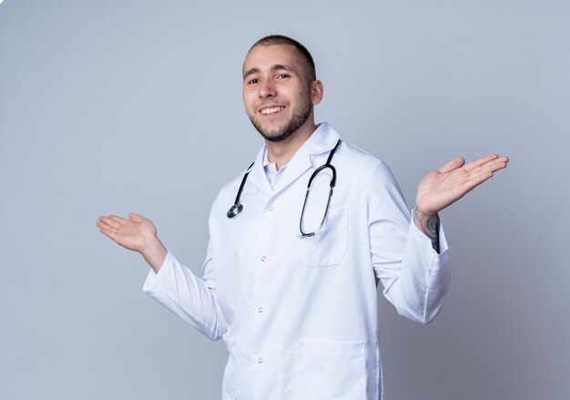 Lächelnder junger männlicher Arzt, der medizinische Robe und Stethoskop um seinen Hals trägt, die leere Hände lokalisiert auf weißer Wand zeigen