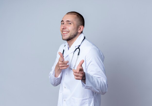 Lächelnder junger männlicher Arzt, der medizinische Robe und Stethoskop um seinen Hals trägt, die Sie Geste an der Front lokalisiert auf weißer Wand tun