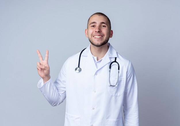 Lächelnder junger männlicher Arzt, der medizinische Robe und Stethoskop um seinen Hals trägt Friedenszeichen lokalisiert auf weißer Wand trägt
