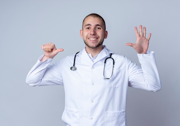 Lächelnder junger männlicher Arzt, der medizinische Robe und Stethoskop um seinen Hals trägt, zeigt sechs mit den Händen lokalisiert auf weißer Wand