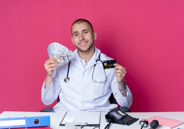 Lächelnder junger männlicher Arzt, der medizinisches Gewand und Stethoskop trägt, sitzt am Schreibtisch mit Arbeitswerkzeugen, die Kreditkarte und Geld lokalisiert auf rosa Wand halten