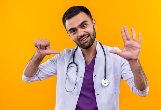 Lächelnder junger männlicher Arzt, der medizinisches Stethoskopkleid trägt, das verschiedene Geste auf lokalisiertem gelbem Hintergrund zeigt