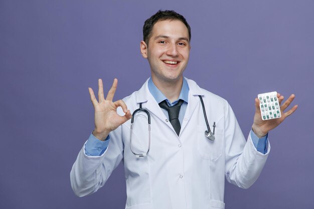Lächelnder junger männlicher Arzt mit medizinischem Gewand und Stethoskop um den Hals, der in die Kamera blickt und eine Packung Kapseln und ein Ok-Zeichen isoliert auf violettem Hintergrund zeigt
