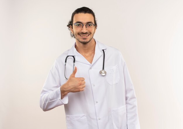 Lächelnder junger männlicher Arzt mit optischer Brille, die weiße Robe mit Stethoskop seinen Daumen oben auf isolierter weißer Wand mit Kopienraum trägt