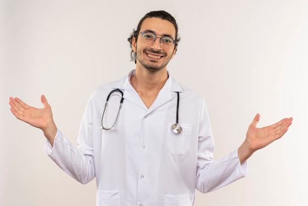 Lächelnder junger männlicher Arzt mit optischer Brille, die weiße Robe mit Stethoskop trägt, breitet Hände auf isolierter weißer Wand mit Kopienraum aus