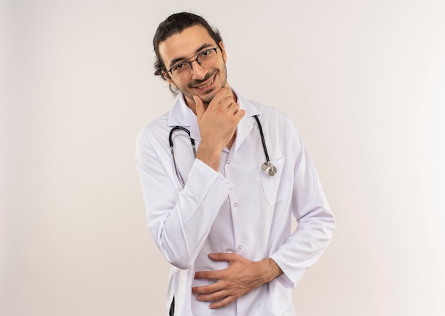 Lächelnder junger männlicher Arzt mit optischer Brille, die weiße Robe mit Stethoskop trägt, die Hand auf Kinn auf isolierter weißer Wand mit Kopienraum setzt