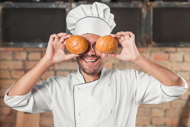 Lächelnder junger männlicher Bäcker, der Brötchen über den Augen hält