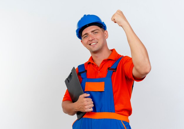 Lächelnder junger männlicher Baumeister, der Uniform und Schutzhelm trägt, Zwischenablage hält und starke Geste auf Weiß tut