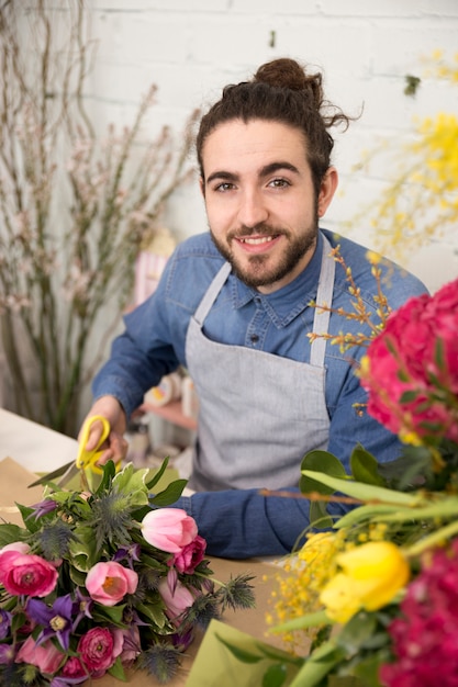 Lächelnder junger männlicher Florist, der die Kamera macht den Blumenblumenstrauß betrachtet